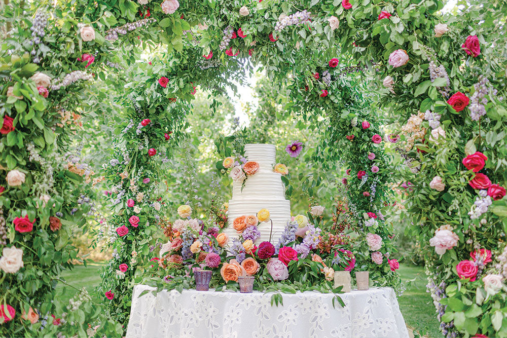 Garden wedding cake table framed by a floral arch of roses and greenery